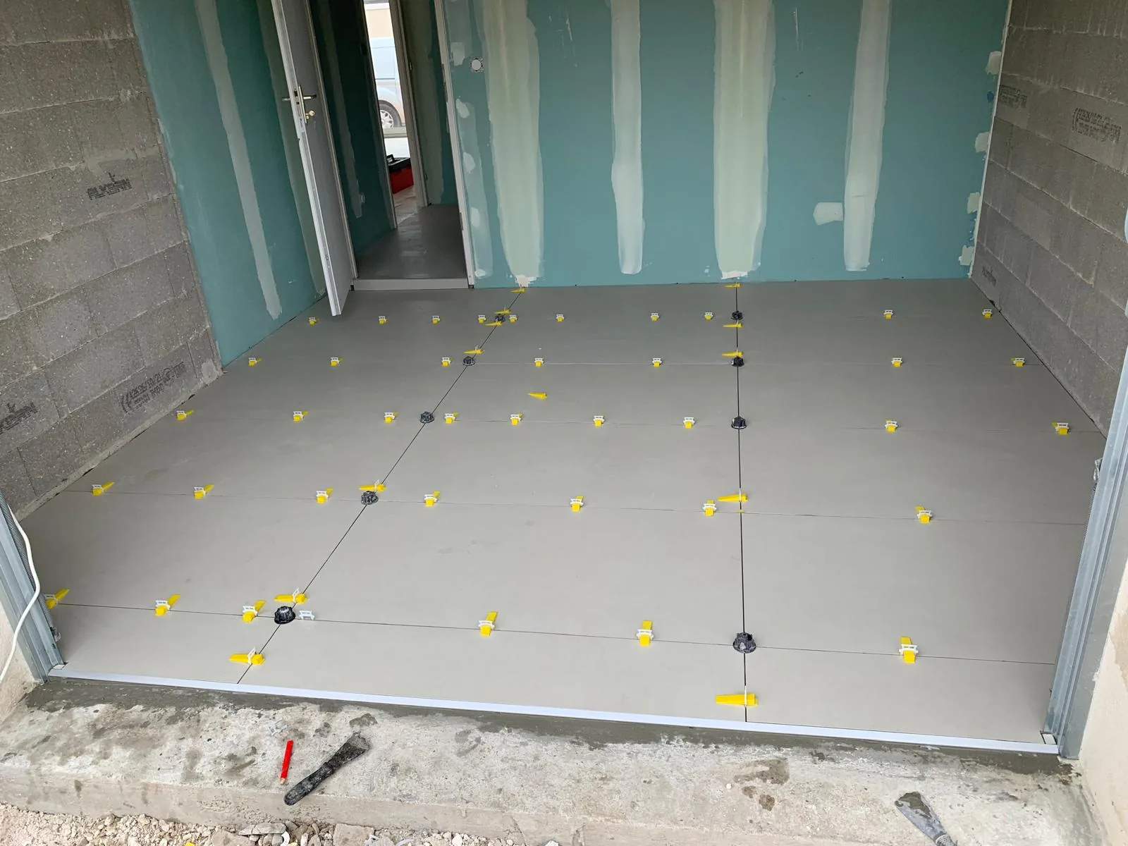 Sol en cours de carrelage grand format effet beton clair, croisillons autonivelants jaunes et plots noirs repartis sur les joints ; murs en plaques de platre hydrofuges vertes et parpaings apparents